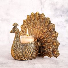 a golden peacock candle holder with a lit candle in front of it on a white surface