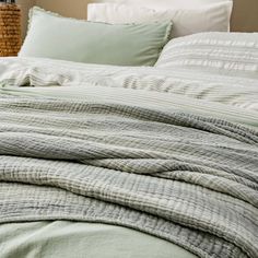 an unmade bed with green linens and pillows on it, in front of a wicker basket