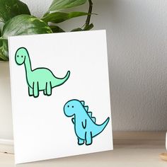 two green and blue dinosaurs on white paper next to a potted plant in front of a white wall