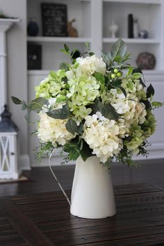 a white vase filled with green and white flowers on top of a wooden table in front of a fireplace