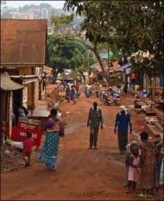 a group of people walking down a dirt road next to buildings and trees in the background