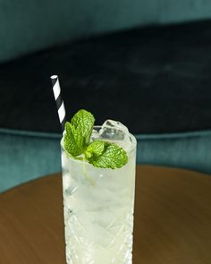 a tall glass filled with ice and mint on top of a wooden table next to a blue velvet chair