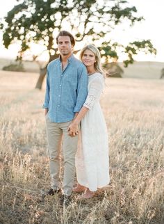 a man and woman holding hands in an open field with a large tree behind them