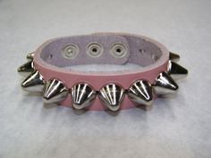 "1 Row Large 1/2\" UK/77 Apex Cone Studs Genuine Leather Handcrafted Bracelet This a handcrafted, genuine leather 3/4\" wide Cuff/bracelet. The Leather is nice and soft, it will get buttery and more subtle with wear. The leather is available in vibrant colors. Racing Red Midnight Black Pretty Pink Arctic White Please check select color at time of check out. The studs/spikes are large UK77 Cone studs. The pyramids studs measure 1/2\" in diameter and conical shaped The Cone studs have a nice lustr Adjustable Punk Wristband With Spikes, Adjustable Spiked Punk Wristband, Adjustable Spiked Bracelets For Festivals, Adjustable Spike Bracelet For Concerts, Adjustable Rock Style Bracelet With Studs, Adjustable Spiked Bracelets For Concerts, Adjustable Silver Wristband With Rivets, Adjustable Edgy Pink Jewelry, Edgy Adjustable Pink Jewelry