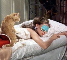 a woman laying in bed with a sleeping mask on her face next to a cat