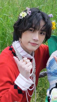 a young man sitting in the grass next to a woman wearing a red sweater and pearl necklace