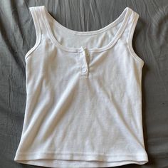 That Tank Top Is A Classic White Color. It's Sleeveless And Looks Really Comfy. The Ribbed Texture Adds A Nice Touch. There's A Single Button Closure In The Front. The Gray Surface Makes The Shirt Stand Out. The White Color Gives A Clean And Fresh Look. The Sleeveless Design Is Perfect For Warm Days. Ribbed Texture Adds A Subtle Detail To The Top. The Button Closure Adds A Stylish Element. It Seems Like A Versatile Piece For Any Outfit. The White Color Is Timeless And Easy To Match. The Ribbed T Sleeveless Cotton Crop Top With Button Closure, Sleeveless Button Closure Crop Top, Casual Sleeveless Crop Top With Button Closure, White Buttoned Tank Top For Summer, Sleeveless Cotton Crop Top With Buttons, Casual White Buttoned Tank Top, White Buttoned Tank Top, White Sleeveless Top With Buttons, Fitted Tank Top With Button Closure