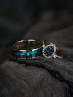 Two unique wedding rings from Staghead Designs sit on a piece of driftwood. These rings are inspired by Aurora Borealis, or the Northern Lights. On the left is a 14K yellow gold wedding band with dark maple wood mountains & an indigo sky made of opal & glow-in-the-dark powder. On the right is a 14K yellow gold "KB" engagement ring with a pear cut lab-created alexandrite center stone & diamond halo & accents. Engraved Wedding Ring, Engraved Wedding Rings, Cute Engagement Rings, Staghead Designs, Future Engagement Rings, Alexandrite Engagement Ring, Engraved Wedding, Men's Wedding Ring, Dream Engagement Rings