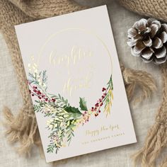 a white card with gold foil and greenery on it next to a pine cone