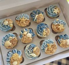 twelve cupcakes in a white box with blue icing and seashells