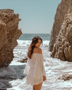 a woman standing on the beach next to some rocks and water with her hair blowing in the wind