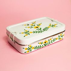 a white lunch box with yellow flowers and green leaves on the lid sitting on a pink surface