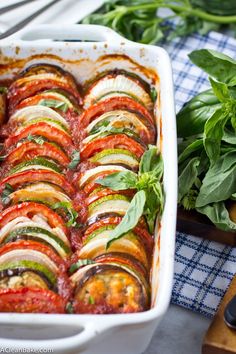 a casserole dish with tomatoes, zucchini and basil on the side