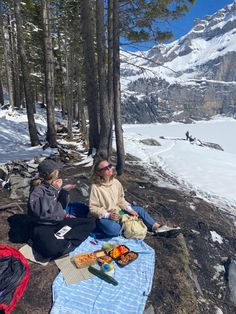 two people sitting on the ground with food and drinks in front of snow covered mountains