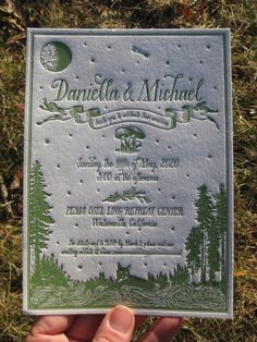 a hand holding up a wedding card in the grass with trees and moon on it