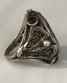 "This lovely filigreed and ornate ring has an oval shaped faceted amethyst purple color glass stone at its center. It is flanked on either side of to and bottom with small round bezel set purple glass stones. The sides have lattice shaped cut outs and the band is smooth. The face of this ring is about 3/4\" long and about 1/2\" across. It is a size 3 1/4 and is marked Sterling. Please note there are some overall signs of wear, especially to the stones, four of five of which, including the center Silver Jeweled Ring For Anniversary, Antique Oval Purple Jewelry, Antique Purple Oval Jewelry, Vintage Jeweled Rings, Ornate Oval Amethyst Ring, Silver Amethyst Ring With Intricate Design, Purple Oval Filigree Rings, Vintage Purple Rings With Accent Stones, Oval Amethyst Ring With Filigree Detail