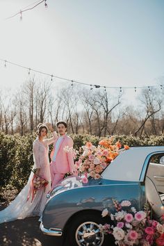 two people standing in front of a car with flowers on the hood and one person wearing a pink dress