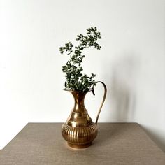 a gold vase with green plants in it on a brown tableclothed surface next to a white wall