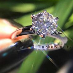 a diamond ring being held by someone's hand with green leaves in the background