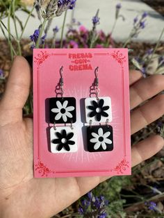 a pair of black and white flower dangle earrings on a pink card with purple flowers in the background