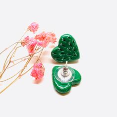 two green heart shaped earrings sitting next to pink flowers on a white surface with one earring in the shape of a leaf