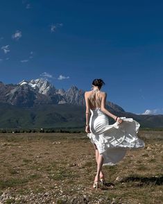 a woman in a white dress is walking through the grass with mountains in the background