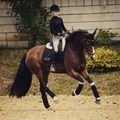 a woman riding on the back of a brown horse across a dirt field in front of a building