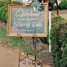 a welcome sign with flowers on it in front of some bushes and shrubbery at a wedding