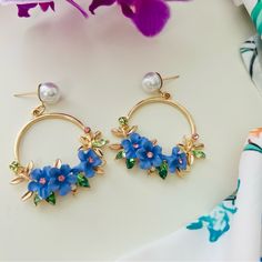 These Stunning Hoop Earrings Showcase A Vibrant Floral Design, With Delicate Blue Flowers Adorned With Pink And Green Crystals. The Golden Hoops Are Accented With Elegant Leaf Details And Are Topped With A Lustrous Pearl Stud, Adding A Touch Of Sophistication. These Earrings Are Perfect For Those Who Love Bold And Colorful Accessories, Making Them A Beautiful Statement Piece For Both Casual And Formal Occasions. Their Playful Yet Refined Style Is Sure To Bring A Fresh And Lively Vibe To Any Outfit. Blue Spring Earrings, Blue Spring Earrings For Pierced Ears, Spring Party Blue Jewelry, Blue Flower-shaped Jewelry For The Beach, Blue Flower Jewelry For The Beach, Elegant Flower-shaped Earrings For Beach, Blue Flower-shaped Spring Jewelry, Floral Print Flower Jewelry For Parties, Blue 3d Flower Drop Earrings