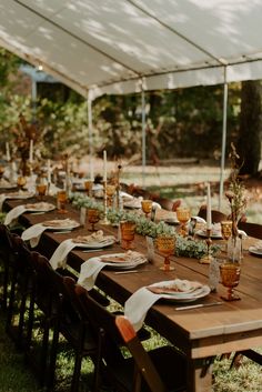 a long table set with place settings for dinner under a tented area in the woods