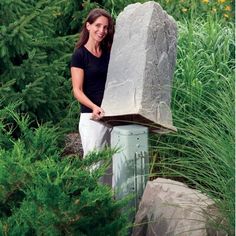 a woman standing next to a large rock in the middle of some bushes and trees