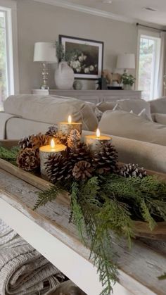 candles are lit on a tray with pine cones and greenery in front of a couch