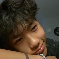 a young man with curly hair wearing a black shirt and smiling at the camera while leaning on his arm