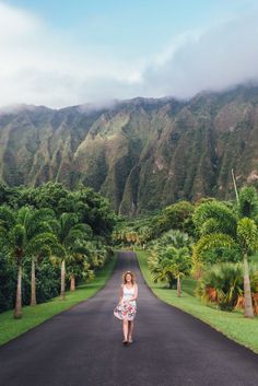 a woman standing in the middle of a road surrounded by palm trees and mountain range