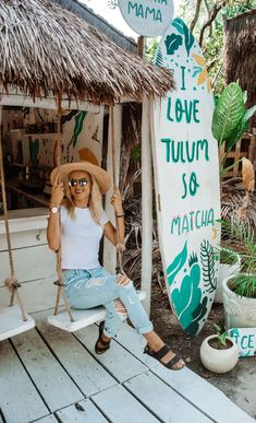 a woman sitting on a swing chair in front of a tiki hut and surfboard