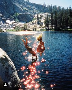 two people jumping into the water from rocks in front of a lake with lights on it