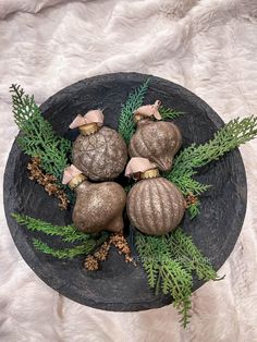 four ornaments are placed on a black plate with greenery and pine cones in the center