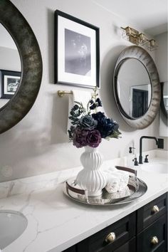 a white vase with flowers sitting on top of a bathroom counter next to two mirrors