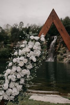 a wedding arch decorated with white flowers and greenery next to a lake in front of a waterfall