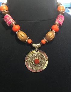 Necklace by Erica Lyons is 12 inches long and adorned with multiple materials; pendant, beads, spacers, chain, and lobster clasp.  Colors include; red, orange, brown, and gold.  Pendant is 2-3/8 inches in diameter with a red stone inlaid.  The necklace weighs 3.5 ounces. Red Metal Jewelry With Colorful Beads, Orange Beaded Amulet Jewelry, Orange Bohemian Beaded Necklaces With Round Shape, Orange Round Bohemian Beaded Necklaces, Orange Bohemian Round Beaded Necklaces, Orange Round Bohemian Beaded Necklace, Red Pendant Beaded Necklace For Festivals, Red Bohemian Necklace With Lobster Clasp, Bohemian Red Necklace With Lobster Clasp