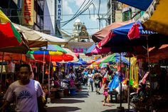 people are walking through an outdoor market with colorful umbrellas on the sides and buildings in the background