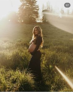 a pregnant woman standing in tall grass with the sun shining down on her belly and trees behind her