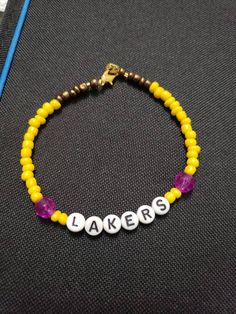 Simple yellow beaded bracelet with purple accent beads and it reads "Lakers" This is unisex adult standard size Personalized Yellow Casual Bracelet, Casual Yellow Personalized Bracelet, Casual Yellow Bracelets With Letter Beads, Personalized Adjustable Yellow Beaded Bracelets, Yellow Friendship Bracelets With Letter Beads, Personalized Adjustable Yellow Friendship Bracelets, Personalized Yellow Friendship Bracelets, Casual Yellow Friendship Bracelets With Letter Beads, Personalized Yellow Bracelets With Round Beads