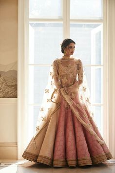 a woman standing in front of a window wearing a pink and gold dress with long sleeves