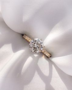 a diamond engagement ring sitting on top of a white satin cloth with the shadow of a flower behind it