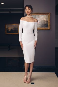 a woman standing in front of a painting wearing a white dress with off the shoulder sleeves