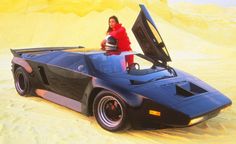 a man standing on top of a black car in the middle of sand dunes with its hood open