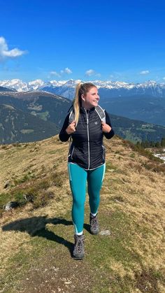 a woman standing on top of a grass covered hillside with mountains in the background,