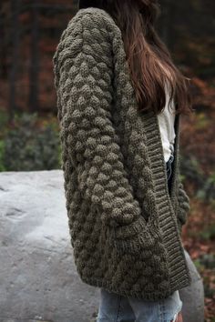 a woman standing in front of a rock wearing a green cardigan sweater and jeans