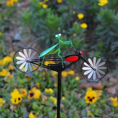 a green frog sitting on top of a wind spinner in front of yellow flowers